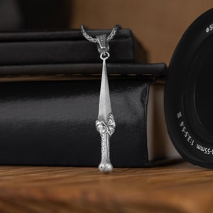 May include: A silver pendant in the shape of a dagger with intricate details. The pendant is hanging from a silver chain and is resting on top of a stack of black books.
