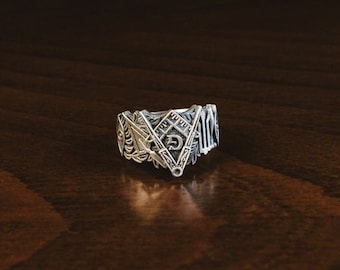 May include: A silver ring featuring detailed geometric and symbolic patterns, including a triangle and the letter "G". The ring is presented on a dark wooden surface, highlighting its intricate design and distinctive style.