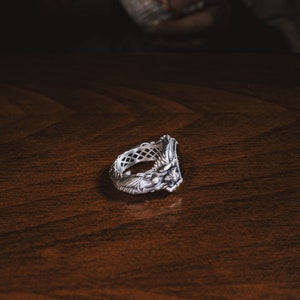 May include: A silver ring with an intricate design featuring a black stone. The ring is on a dark wood surface.