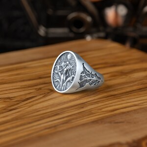 May include: A silver ring with a detailed design of a woman standing in a forest with a full moon in the background. The ring is oval shaped and has a tree branch design on the side.