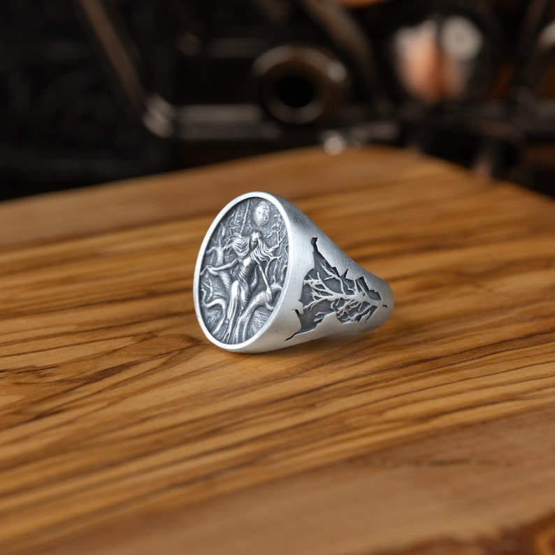 May include: A silver ring with a detailed design of a woman standing in a forest with a full moon in the background. The ring is oval shaped and has a tree branch design on the side.