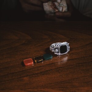 May include: A silver ring with a black gemstone set in a square shape. The ring is surrounded by four smaller gemstones: red, brown, and two green.