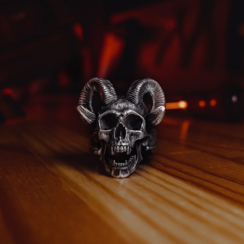 May include: A silver skull ring with horns, the skull has an open mouth with teeth and a detailed design.