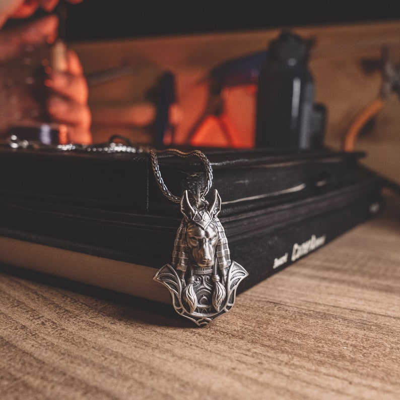 May include: A silver pendant necklace with a detailed Anubis head design. The pendant is resting on a stack of black books.