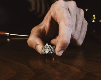 Puede incluir: Un anillo plateado con símbolos masónicos, incluyendo una escuadra y compás con la letra "G" en el centro. El anillo es sostenido por una mano, mostrando sus intrincados detalles. El anillo está sobre una superficie de madera.