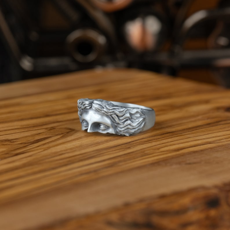 Op de afbeelding: Een zilveren ring met een gedetailleerd gesculpteerd gezicht van een vrouw met golvend haar. De ring ligt op een houten oppervlak.