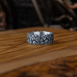 May include: A silver ring with a detailed design of swirling clouds. The ring is wide and has a smooth, polished finish.