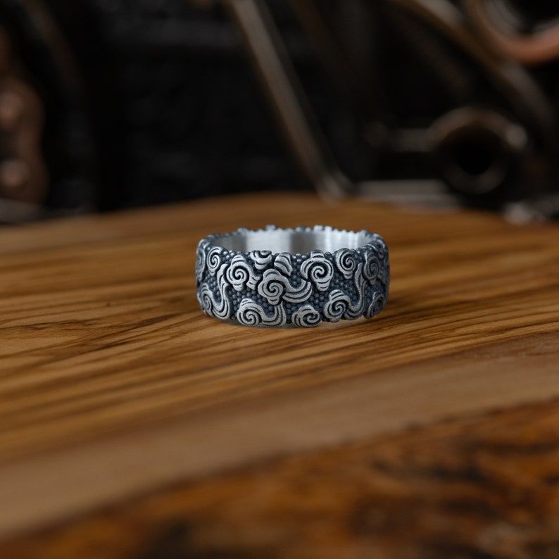 May include: A silver ring with a detailed design of swirling clouds. The ring is wide and has a smooth, polished finish.
