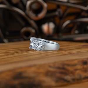 Op de afbeelding: Een zilveren ring met een gedetailleerd gezichtontwerp. De ring ligt op een houten oppervlak.