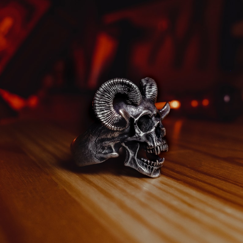 May include: A silver skull ring with horns, the skull has an open mouth and detailed features.