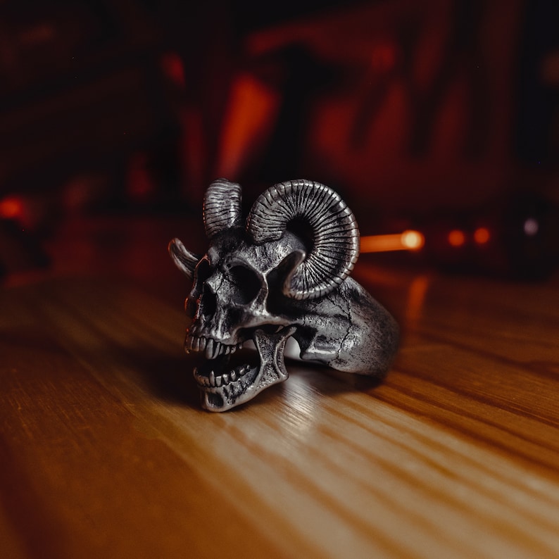May include: A silver skull ring with ram horns. The ring is detailed with a skull face and horns.