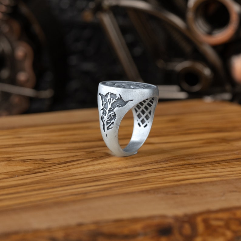 May include: A silver ring with a detailed tree design on the face. The ring has a textured band.