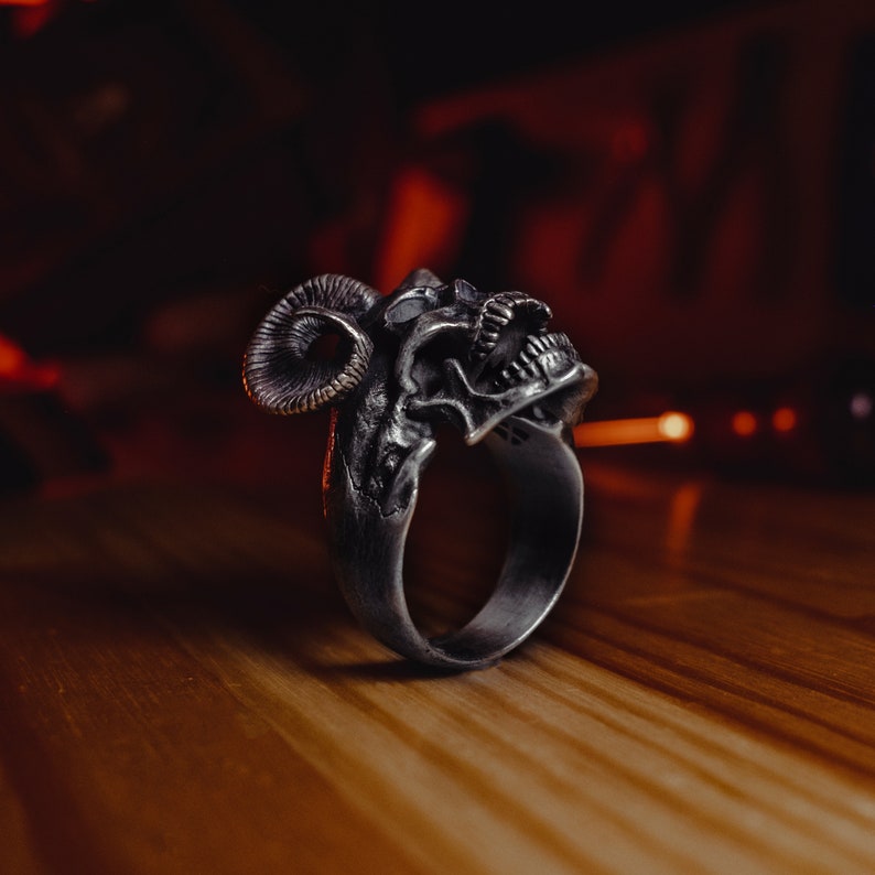 May include: A silver ring with a detailed skull design and ram horns. The ring is on a wooden surface.