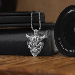 May include: Silver necklace with a detailed, three-dimensional pendant of a Japanese demon mask. The pendant has a textured surface and intricate details.
