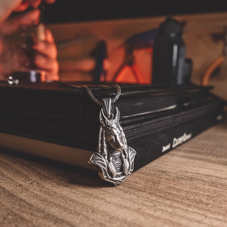 May include: A silver pendant necklace with a detailed Anubis head design. The pendant is resting on a stack of black books.
