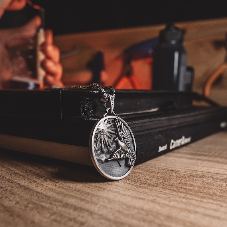 May include: A silver pendant necklace with a round, detailed design of a bird in flight. The pendant is resting on top of two black books.