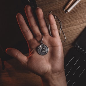 May include: A silver necklace with a round pendant featuring a stylized bird design. The pendant is held in a person's hand.