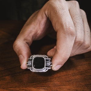 May include: A silver ring with a black stone set in a square frame. The ring has intricate details and a textured finish.