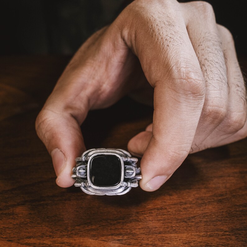 May include: A silver ring with a black stone set in a square frame. The ring has intricate details and a textured finish.