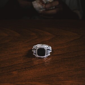 May include: A silver ring with a black gemstone set in a square shape. The ring has intricate details and a textured band.