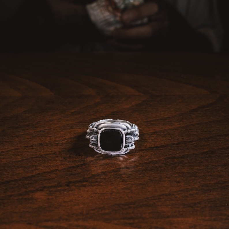 May include: A silver ring with a black gemstone set in a square shape. The ring has intricate details and a textured band.