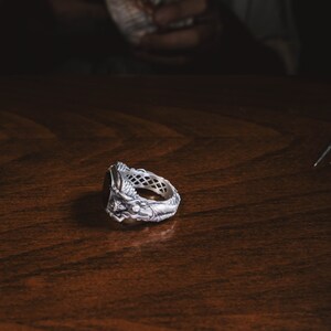 May include: A silver ring with a detailed design featuring a figure and a black stone. The ring is lying on a wooden surface.