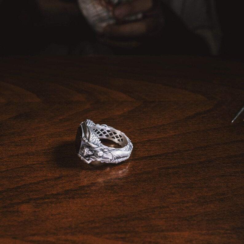 May include: A silver ring with a detailed design featuring a figure and a black stone. The ring is lying on a wooden surface.