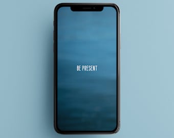 以下が含まれることがあります： 青いぼかしの背景に「BE PRESENT」というテキストが表示された黒いスマートフォン