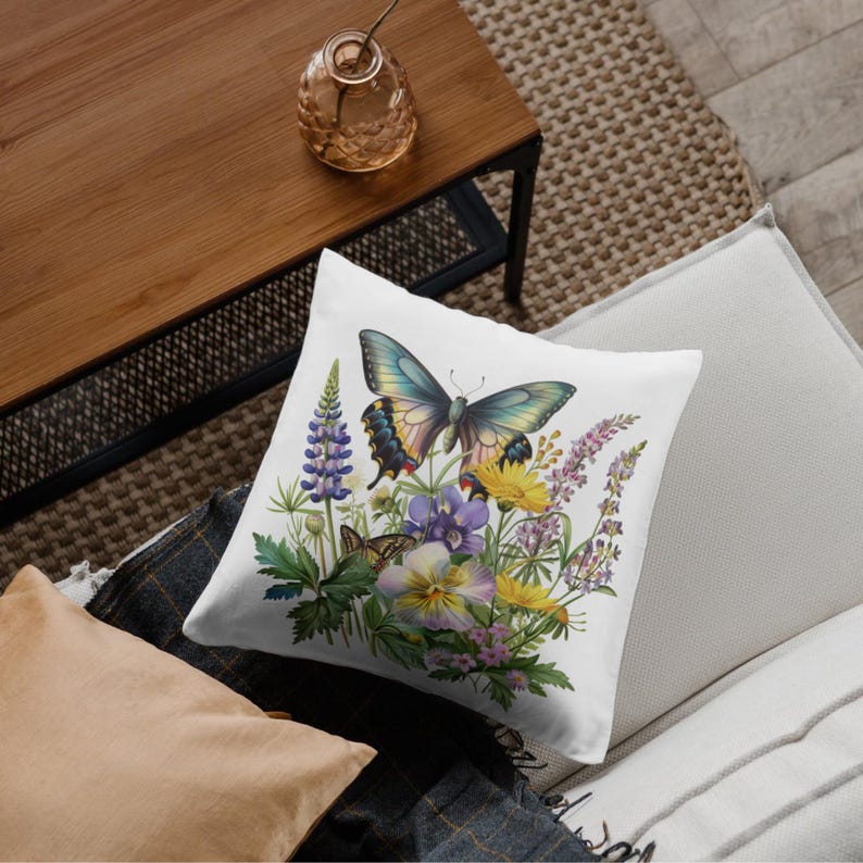 Puede incluir: Coj&iacute;n cuadrado blanco con un dise&ntilde;o de mariposa y flores. El coj&iacute;n presenta una gran mariposa con alas azules, amarillas y negras, rodeada de flores moradas, amarillas y verdes. El coj&iacute;n est&aacute; sobre un sof&aacute;.