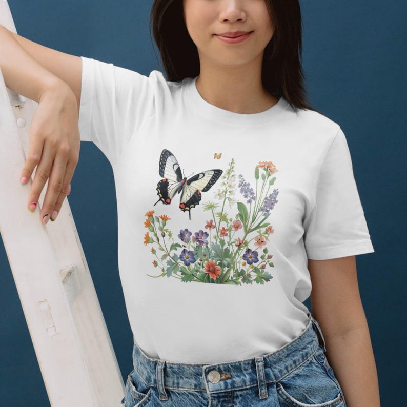 Puede incluir: Camiseta blanca con un dise&ntilde;o de mariposa y flores. La mariposa tiene alas negras y blancas, y las flores son en tonos morados, naranjas y blancos. La camiseta es de material suave.