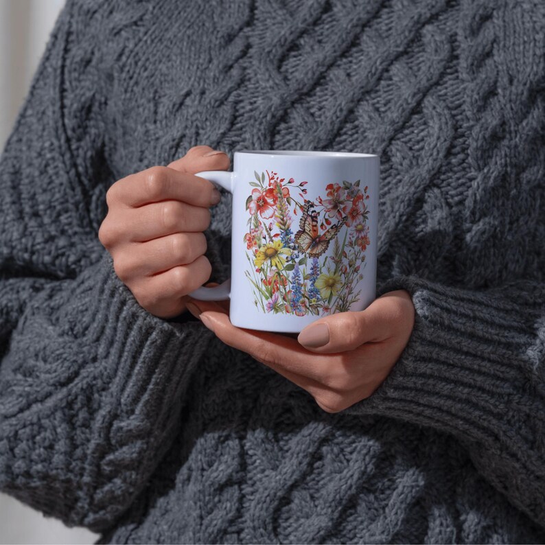 Puede incluir: Una taza de cer&aacute;mica blanca con un colorido dise&ntilde;o floral y de mariposas. El dise&ntilde;o incluye flores rojas, amarillas y azules con una mariposa en el centro. La taza tiene un asa blanca y la sostiene una persona con un su&eacute;ter gris.