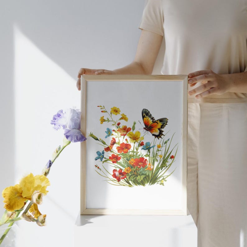 Puede incluir: Una impresi&oacute;n bot&aacute;nica enmarcada con una colorida ilustraci&oacute;n de flores silvestres y una mariposa. La obra de arte muestra flores rojas, amarillas y azules con una mariposa detallada, todo sobre un fondo blanco dentro de un marco de madera clara.