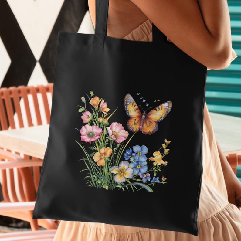 Puede incluir: Bolso tote negro con un dise&ntilde;o floral y de mariposa colorido. La ilustraci&oacute;n incluye flores rosas, naranjas, amarillas y azules con una mariposa naranja detallada. El bolso tiene una forma rectangular simple y un asa.
