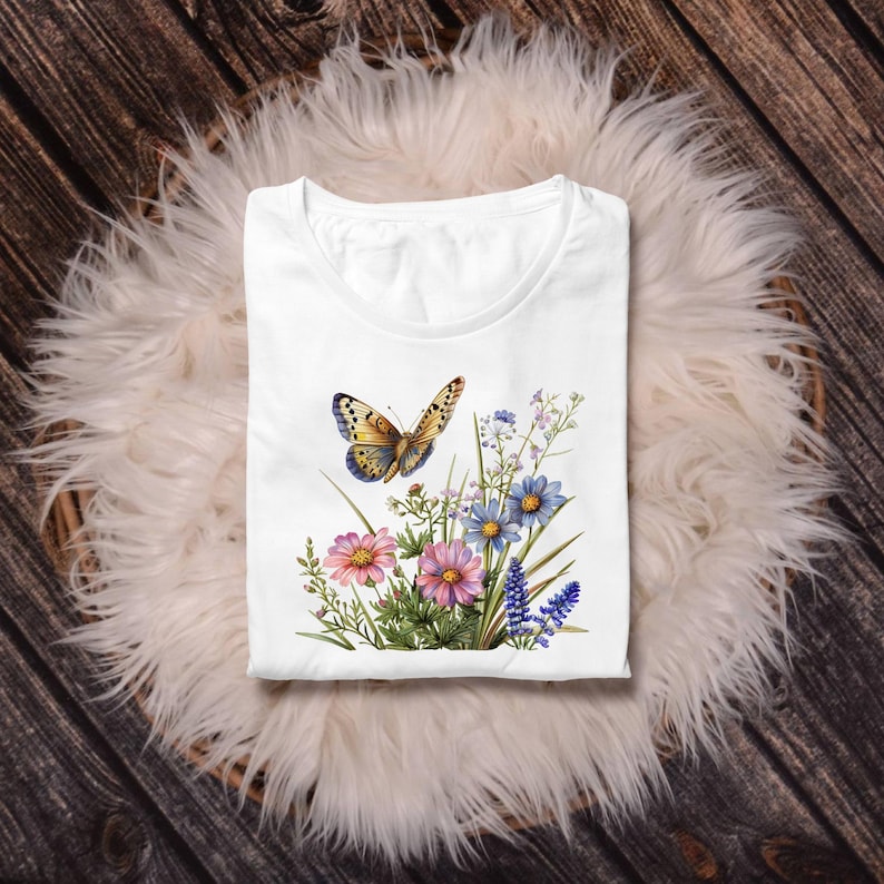 Puede incluir: Camiseta blanca con un dise&ntilde;o de mariposa y flores. El gr&aacute;fico incluye flores rosas, azules y moradas con hojas verdes. La mariposa tiene alas amarillas y azules. La camiseta est&aacute; doblada sobre una superficie blanca y esponjosa.