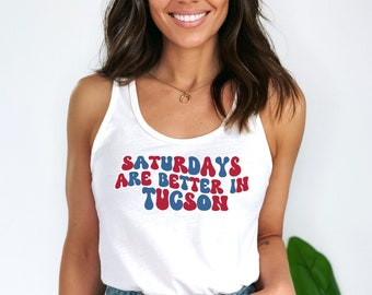 May include: A white tank top with the text "Saturdays are better in Tucson" in red and blue lettering. The text is in a retro font style.
