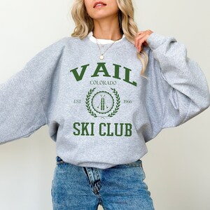 May include: A gray sweatshirt with the text "VAIL COLORADO SKI CLUB" in green. The design includes a circular emblem with crossed skis and a laurel wreath. The sweatshirt has a crew neck and long sleeves.