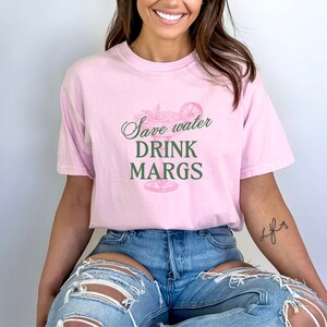 May include: A pink t-shirt with the text "Save water drink margs" and a green illustration of a margarita glass with a lime wedge.