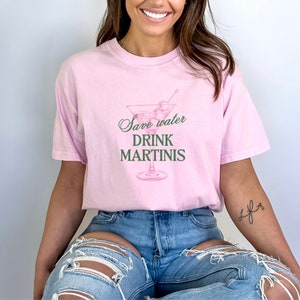 May include: A pink t-shirt with the text "Save water drink martinis" and a pink martini glass with green olives.