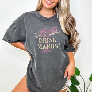 May include: A dark gray t-shirt with a pink margarita glass graphic and the text "Save water drink margs" in a gold script font.