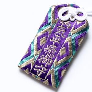 May include: An embroidered Japanese omamori charm in purple, teal, and gold. The charm has a geometric pattern and Japanese characters. A white cord and knot are attached to the top. The charm is rectangular.