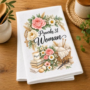 Könnte beinhalten: Ein weißes Waffelpiqué-Küchentuch mit einem Blumenkranz, einem Holzkreuz mit Perlen, einem Stapel Bücher mit einer Tasse mit der Aufschrift blessed sowie einem Korb mit Garn und einer Schere. Der Text Proverbs 31 Woman ist in Schwarz gedruckt.
