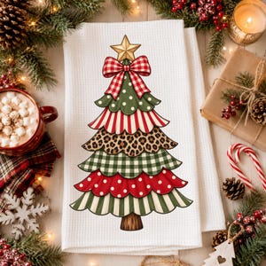 Pode incluir: Um pano de prato branco com textura de waffle apresentando um gráfico central de uma árvore de Natal com camadas recortadas em padrões vermelhos e verdes, incluindo bolinhas, listras e estampa de leopardo, com uma estrela dourada e laço.