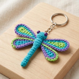 May include: A crocheted dragonfly keychain featuring a bright blue body and two purple eyes. The four wings are layered with a light green center, a teal middle section, and a purple outer border, all attached to a silver metal split ring.