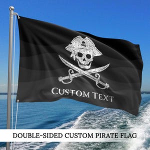 May include: Black double-sided custom pirate flag with a white skull and crossbones design, pirate hat, and crossed swords. The words "CUSTOM TEXT" are printed below the skull. The flag is waving in the wind against a blue sky and water background.