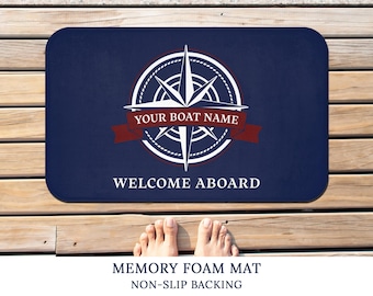 以下が含まれることがあります： 白いコンパスデザインと「YOUR BOAT NAME」と「WELCOME ABOARD」のテキストが書かれた青いウェルカムマット。マットはメモリーフォーム製で、滑り止め加工が施されています。