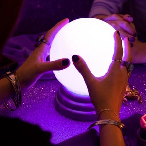 Pode incluir: Uma bola de cristal branca brilhante, segurada por mãos adornadas com anéis e pulseiras, contra um fundo roxo brilhante. A bola é iluminada, criando um ambiente místico. A cena sugere adivinhação ou clarividência.