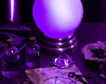 May include: A glowing purple crystal ball sits on a metallic stand, surrounded by mystical items. Two glass bottles with cork stoppers, a large gem, a feather, a key, and a pentagram drawing complete the scene. The items are arranged on a dark surface.