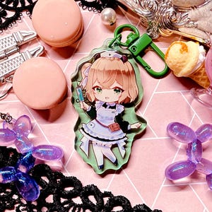 May include: A clear acrylic keychain featuring an anime-style nurse character holding a syringe. The character has blonde hair, green eyes, and wears a white and blue uniform. The keychain has a green clasp. The background includes pink macarons and decorative elements.