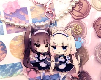 Könnte beinhalten: Ein klarer Acryl-Anhänger mit zwei Anime-Charakteren mit Katzenohren, die Maid-Outfits tragen. Der Anhänger ist an einem herzförmigen rosafarbenen Verschluss befestigt. Eine Perlenkette und dekorative Wachssiegel sind ebenfalls zu sehen.