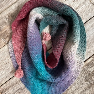 May include: A hand-knitted shawl in a gradient of colors, including teal, purple, and burgundy. The shawl has a triangular shape and is adorned with two small tassels. The soft, textured fabric is displayed on a rustic wooden surface.